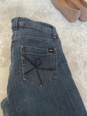 Mörkblå Vintage bootcut jeans med broderad ficka - Säljer ett par mörkblå bootcut jeans med snygg broderad detalj på bakfickan. Jeansen har klassisk femficksmodell, bälteshällor och subtila slitningar. Perfekta för dig som gillar en avslappnad men ändå trendig look.