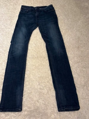 Nudie Jeans - Snygga mörkblå jeans med rak passform och klassisk femficksdesign. Jeansen har subtila slitningar och kontrastsömmar. Tillverkade i denim med en tidlös look som passar till det mesta. Perfekta för dig som gillar enkel och stilren stil.