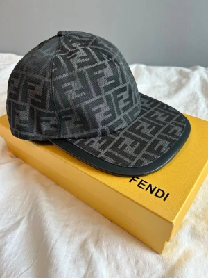 Svart keps med Fendi-logga - Snygg svart keps från Fendi med ikoniskt FF-monogram i grått över hela kepsen. Klassisk böjd skärm med svart kant i läderlook och justerbar baksida. Perfekt accessoar för dig som gillar exklusiv streetwear och vill sticka ut.