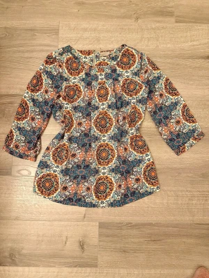 Mönstrad blus från Vero Moda XS - Färgglad blus från Vero Moda i storlek XS med ett bohemiskt mönster i blått, orange, vitt och gult. Blusen har trekvartsärm och rund halsringning samt dragkedja bak. Perfekt för dig som gillar unika prints och en avslappnad stil.