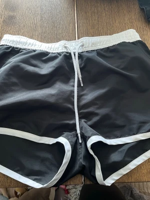Svarta badbyxor från Frank Dandy - Snygga svarta badbyxor från Frank Dandy med vita detaljer och dragsko i midjan. De har vit resår och meshfoder på insidan för extra komfort. Perfekta för stranden eller poolen.