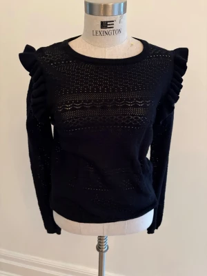 Svart stickad tröja med volang - Snygg svart stickad tröja från & Other Stories i storlek S (36-38). Tröjan har volangdetaljer på axlarna och ett genombrutet mönster över hela plagget. Rund halsringning och långa ärmar ger en trendig och cool look.