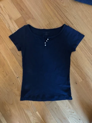 Mörkblå topp med knappar - En mörkblå, tight topp med korta ärmar och rundad halsringning. Toppen har tre små knappar framtill som detalj och är tillverkad i ett mjukt bomullsmaterial. Perfekt att styla med jeans eller kjol för en enkel och snygg look. Använd max 3 gånger.💓