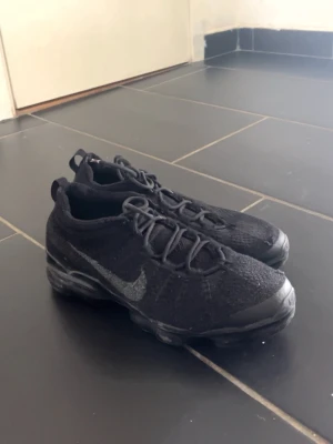 Nike svarta vapor Max - Säljer ett par helsvarta Nike sneakers med Flyknit-överdel och synlig Air-sula. Skorna har en låg profil, rund tå och diskret Nike-logga på sidan. Perfekta för dig som gillar stilrena och sportiga skor med skön passform.