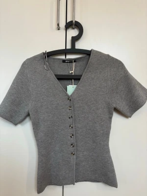 Grå topp med knappar - Snygg grå ribbad topp från Gina Tricot med v-ringning och korta ärmar. Toppen har dekorativa knappar framtill och en figurnära passform som framhäver siluetten. Perfekt att styla med jeans eller kjol.