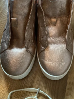 Beige/ Roséguld sneakers från Bugatti  - Snygga sneakers i metallic roséguld från Bagheera. Skorna har en låg profil, vita sulor och är gjorda i ett glansigt syntetmaterial. Perfekta för dig som vill sticka ut med en unik färg på fötterna. Klassisk design utan snören på bilderna.