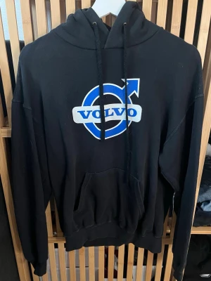 Svart Volvo hoodie med tryck - Svart hoodie med stor Volvo-logga i blått och vitt på bröstet och ett k rollat thext-tryck på ryggen. Klassisk känguruficka framtill, huva med dragsko och ribbade muddar. Perfekt för dig som vill sticka ut och visa din Volvo-pride.