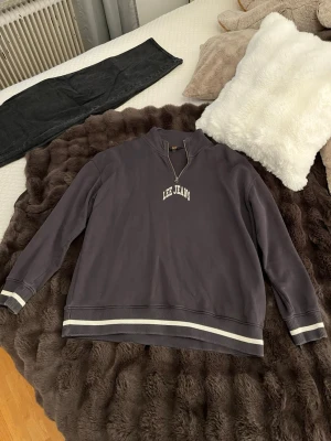 Lee Jeans quarterzip i mörkgrå - Mörkgrå hoodie från Lee Jeans med vit logga på bröstet. Tröjan har halv dragkedja vid halsen, ribbade muddar och nederkant med vita ränder. Storlek L