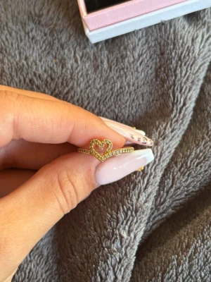 Guldfärgad ring med hjärta - Söt guldfärgad ring med ett öppet hjärta i mitten, dekorerad med små gnistrande stenar runt hela hjärtat och längs ringen. Pris kan diskuteras, inge täcken på användning. 