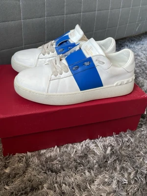 Valentino Garavani vita sneakers - Säljer ett par skit feta vita sneakers från Valentino Garavani perfekt för sommaren med en bred blå rem över snörningen. Skorna har klass profill och sopren design med diskret logga på plösen. Tillverkade i släta material med gummisula och vita skosnören.