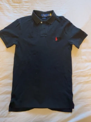 Svart pikétröja från Ralph Lauren - Klassisk svart pikétröja från Ralph Lauren med röd broderad logga på bröstet. Tröjan har korta calendar, krage och knappar framtill. Perfekt till jeans eller shorts för en clean och stilren look.