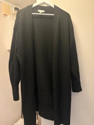 Svart lång kofta i oversized modell - Säljer en svart lång kofta i oversized modell med öppen front och långa ärmar. Koftan är mjuk och har en lite grovstickad struktur, perfekt att slänga över en t-shirt eller hoodie. Enkel och stilren design utan knappar eller dragkedja.