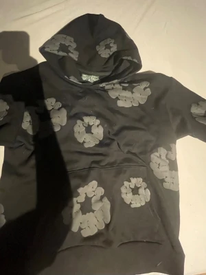 Svart grå denim ters hoodie  - Svart hoodie från Market med stora gråa blommönster över hela plagget. Tröjan har en klassisk huva och en stor magficka framtill. Materialet är mjukt och bekvämt, perfekt för dig som gillar en unik och trendig stil.