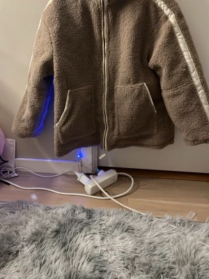 Brun teddyjacka med vit rand - Mysig brun teddyjacka med dragkedja framtill och två stora fickor. Jackan har en vit rand längs ärmarna och vita detaljer vid fickorna. Tillverkad i fluffigt teddy-material som håller dig varm och ger en avslappnad look.