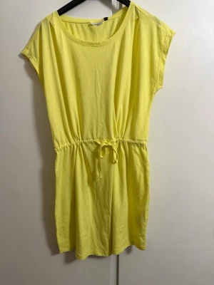 Gul kortklänning med knyt i midjan - En enkel och fräsch gul kortklänning i mjuk bomull. Klänningen har rund halsringning, korta ärmar och ett knytband i midjan för en snygg passform. Perfekt för varma dagar och sommarens alla tillfällen.