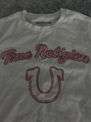 True Religion topp - Snygg vit topp från True Religion med glittriga röda paljetter som formar loggan och hästsko på bröstet. Klassisk rund halsringning och korta ärmar. Perfekt för dig som vill sticka ut med en ikonisk streetwear-look.