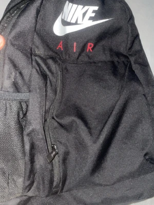 Svart Nike Air ryggsäck - Cool svart ryggsäck från Nike Air med klassisk vit Nike-logga framtill och röd text på axelremmen. Ryggsäcken har en enkel, sportig design med justerbara remmar och slitstarkt material. Perfekt för skola, träning eller utflykter.