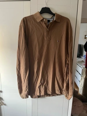 Brun långärmad pikétröja Tommy Hilfiger - Brun långärmad pikétröja från Tommy Hilfiger med klassisk krage och knappar vid halsen. Tröjan har en liten diskret logga på bröstet och är gjord i mjukt material som känns skönt mot huden. Enkel och stilren design som funkar till det mesta.