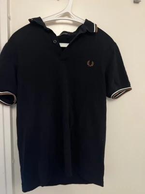 Svart pikétröja från Fred Perry - Klassisk svart pikétröja från Fred Perry med vita och beige detaljer på krage och ärmslut. Tröjan har korta ärmar och den ikoniska Fred Perry-loggan broderad på bröstet. Perfekt till jeans eller shorts för en clean look.