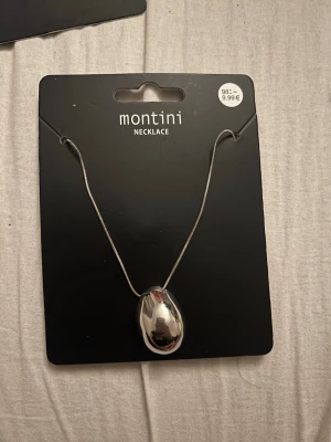 Silverfärgat halsband från Montini - Snyggt halsband från Montini med en stor, droppformad silverfärgad berlock. Kedjan är tunn och smidig, vilket ger ett stilrent och modernt intryck. Perfekt accessoar för att lyfta din outfit med en cool metallisk touch.
