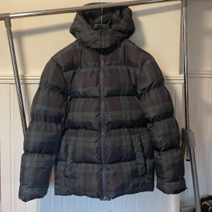 Mörkgrön rutig pufferjacka med huva - Snygg pufferjacka i mörkgrönt och svart rutigt mönster. Jackan har en rejäl huva, dragkedja framtill och två stora fickor. Perfekt för kalla dagar med sin vadderade och puffiga design. Passar dig som vill ha en stilren och varm vinterjacka. Pris kan diskuteras vid snabb affär.