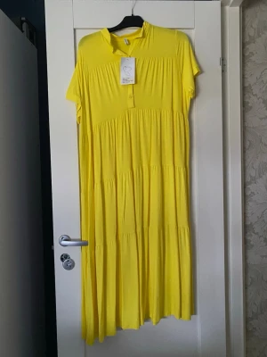 Gul långklänning med kort ärm från Hängmatta - Lång gul klänning med korta ärmar och knappar framtill. Klänningen har en lös passform och är tillverkad i ett mjukt, lätt material som faller fint. Perfekt för varma dagar och har en enkel, stilren design.