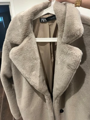 Beige fuskpäls kappa från Zara - Snygg beige kappa från Zara i mjuk fuskpäls med bred krage och knäppning framtill. Kappan har en rak passform och är helfodrad för extra komfort. Perfekt för dig som vill ha en trendig och mysig look under kalla dagar.