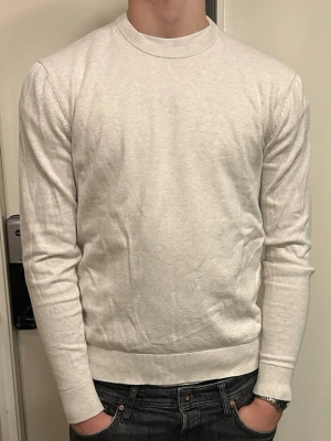 Gräddfärgad tjocktröja - Säljer en stilren gräddfärgad stickad tröja med rund halsringning och långa ärmar. Tröjan har en smal passform och ribbade muddar vid ärmslut och nederkant. Perfekt för en clean och enkel look.