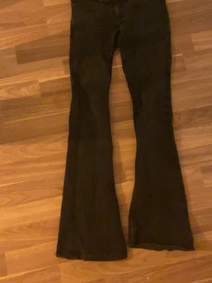 Svarta jeans från gina 14+ - Säljer ett par svarta utsvängda jeans med snygga dekorationer på bakfickorna. Jeansen har låg midja och klassisk femficksdesign. Materialet är denim och passformen är figurnära upptill med breda ben. Perfekta för dig som gillar retrostil och vill sticka ut. De har blivit lite missfärgade i tvätten🫰🏼