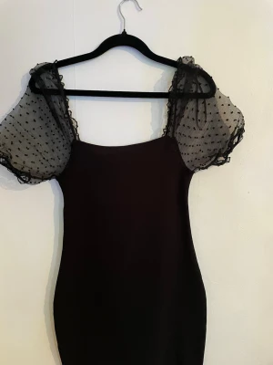 Svart klänning med puffärmar - Säljer en svart klänning med transparenta puffärmar i mesh med små prickar. Klänningen har en figurnära passform och raka linjer, perfekt för dig som vill ha en stilren look med en twist. Ärmarna ger en cool och trendig känsla.