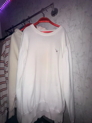 Vit stickad tröja från GANT - Vit stickad tröja från GANT med rund halsringning och diskret broderad logga på bröstet. Tröjan har en klassisk passform och ribbade muddar vid ärmslut och nederkant. Perfekt för en clean och stilren look.