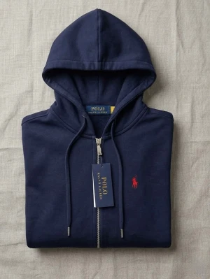 Polo Ralph Lauren | Mörkblå hoodie med dragkedja - mörkblå hoodie från Polo Ralph Lauren med dragkedja, huva med dragsko och klassisk röd logga broderad på bröstet. Mycket bra skick, knappt använt. Med prislapp på.