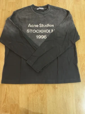 Svart långärmad topp Acne Studios - Cool svart långärmad topp från Acne Studios med vit text 'Acne Studios STOCKHOLM 1996' på bröstet. Klassisk rund halsringning och relaxed fit. Perfekt för dig som gillar stilrena och trendiga plagg med statement-tryck.