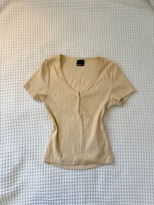 Beige ribbad topp från Gina Tricot - Snygg beige ribbad topp från Gina Tricot med v-ringning och knappar framtill. Modellen är figurnära med korta ärmar och mjukt stretchigt material som sitter skönt på kroppen. Perfekt att styla med jeans eller kjol för en enkel look.