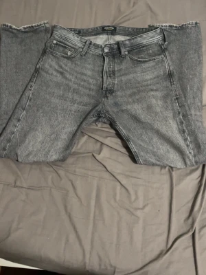 Grå raka jeans från Jack & Jones - Säljer ett par grå jeans från Jack & Jones med rak passform och klassisk femficksdesign. Jeansen har en snygg tvättad look och är tillverkade i denim. Perfekta för en avslappnad och trendig stil.