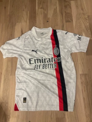 AC Milan bortatröja Puma Rafa Leão - Snygg vit AC Milan bortatröja från Puma med Rafa Leão och nummer 17 på ryggen. Tröjan har svarta och röda ränder på framsidan, klubbmärke på bröstet och sponsortryck. Tillverkad i lätt och ventilerande material, perfekt för fotboll eller träning. Pris kan diskuteras 