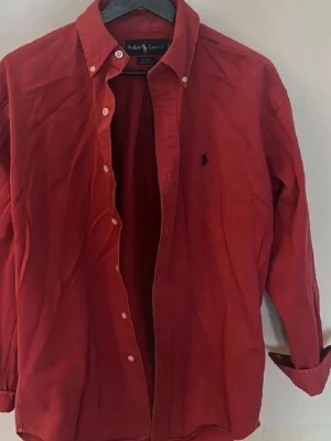 Röd skjorta från Ralph Lauren - Klassisk röd skjorta från Ralph Lauren med button-down krage och broderad logga på bröstet. sopassad passform och långa ärmar. Tillverkad i mjuk bomull och stängs med vita knappar framtill. Perfekt för en stilren och avslappnad look.