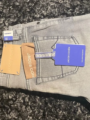 Jacob Cohën jeans i två färger - Två par snygga jeans från Jacob Cohën. Ena paret är ljusgrå med klassisk femficksdesign och detaljerade sömmar, det andra paret är blå denim med kontrastsömmar och unik fickdetalj. Båda har exklusiva etiketter och är tillverkade i mjukt bomullsmaterial.