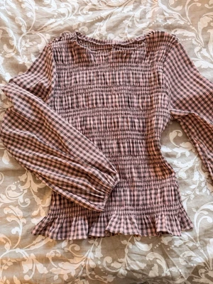 Rutig smockad blus med volangkant - Söt blus i rosa och brunrutigt med smockad kropp och volangkant nedtill. Blusen har långa puffiga ärmar och rund halsringning. Perfekt för dig som gillar romantisk och trendig stil.