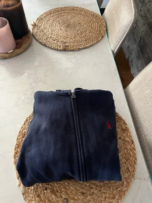 Mörkblå hoodie från Ralph Lauren - Mörkblå hoodie integrerad med dragkedja från Ralph Lauren. Klassisk design med huva, kängurufickor och röd broderad logga på bröstet. Tillverkad i mj composit bomullsmix som känns skön mot huden. Perfekt för en avsl sopad look. Stl: 14 år