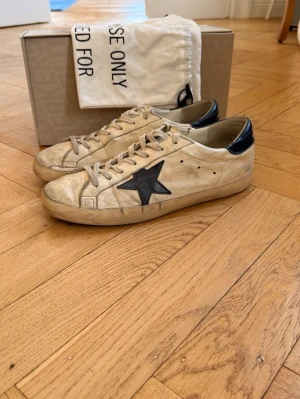 Golden Goose skor - Golden Goose Superstar sneakers i ljus beige läder med svart stjärndetalj på sidan och svart h thread på hälen. Klassisk låg modell med slitna detaljer. Kommer med originalkartong och dustbag. Nypris 5500kr. Bra skick men lite slitningar i sulan, men något man inte ser vid användning. Pris går att diskutera. Hör av er vid frågor och funderingar