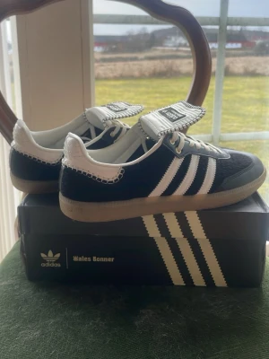 Adidas x Wales Bonner svart/vit sneaker - Unika Adidas x Wales Bonner sneakers i svart mocka och läder med vita detaljer och klassiska tre ränder. Skorna har en bred vit plös med kontrastsömmar och logus monogram. Ytters listing i gummi och vita skosnören. Snygg retrovibe och riktigt statement.