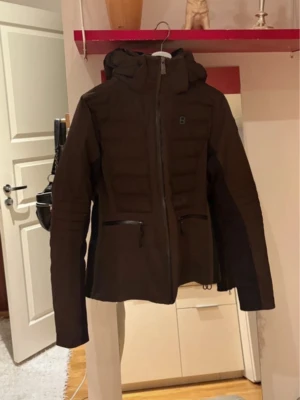 Brun dunjacka med huva från 8848 - Säljer en svart dunjacka från 8848 med huva och dragkedja framtill. Jackan har två fickor med dragkedja och quiltade partier på bröstet. Perfekt för kalla dagar och har en sportig, stilren look.