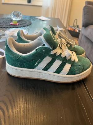 Adidas Campus gröna sneakers - Gröna Adidas Campus sneakers i mocka med vita klassiska ränder på sidorna och vit sula. Skorna har tjocka vita skosnören och Adidas-logga på plösen. Låg modell som passar perfekt till streetwear och avslappnad stil.