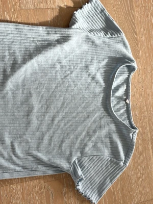 Ljusblå ribbad t-shirt - Säljer en ljusblå ribbad t-shirt med korta ärmar och vågig kant nertill. T-shirten har en enkel, avslappnad passform och är tillverkad i mjuk bomull. Perfekt till jeans eller kjol för en chill look.