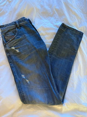  demiblå slitna jeans från Diesel - Säljer ett par klassiska demiblå jeans från Diesel med slitningar på framsidan. Dom är i mycket bra skick nästan sprillans