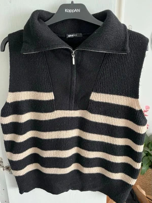 Randig stickad väst från Gina Tricot - Säljer denna snygga stickade västen från Gina Tricot i svart och beige med breda ränder. Västen har en hög krage med half zip-dragkedja. 