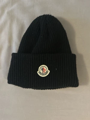 Svart ribbstickad mössa Moncler - Säljer en svart ribbstickad mössa från Moncler med uppvikt kant och klassisk Moncler-logga framtill. Mössan är gjord i ett mjukt och stretchigt material som sitter skönt på huvudet. Perfekt för dig som vill ha en stilren och trendig accessoar.