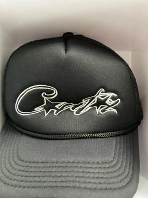 Svart truckerkeps med broderad text - Snygg svart truckerkeps med vit broderad text och stjärnor framtill. Kepsen har mesh på baksidan för extra ventilation och justerbar snapback-spänne. Perfekt för en streetwear-look.