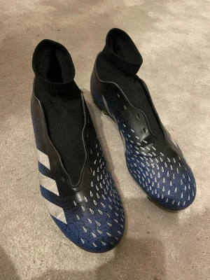 Adidas predator freak fotbollsskor - Säljer ett par Adidas predator freak med hög strumpa. Skorna är svarta, blå och grön med vita detaljer och mönster på ovandelen samt de klassiska tre ränderna på sidan. Ovandelen är i syntetmaterial och designen är modern och sportig. Använda max 5 gånger och svarta snören ingår. 